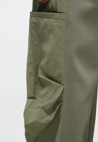 Παντελόνια σε ελιά πράσινο χρώμα, φτιαγμένα από υφασμάτινη υφή, με μια μεγάλη πλαϊνή τσέπη και ένα λείο, ριγέ διακοσμητικό κατά μήκος της πλαϊνής ραφής.