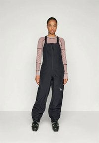 Črne vodoodporne smučarske hlače z nastavljivimi trakovi, sprednjim zadrgo in logotipom. Oblečene nad vzorčasto dolgosleeveno majico in smučarskimi čevlji.