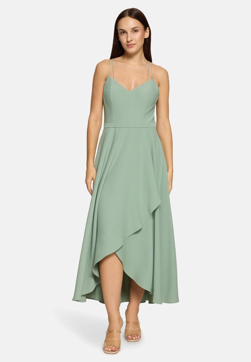 Kobieta w asymetrycznej midi sukience bez rękawów w kolorze szałwii z cienkimi ramiączkami, w beżowych sandałach na obcasie z odkrytymi palcami, stojąca na białym tle.