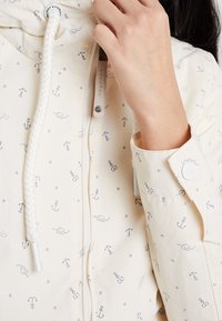 Närbild av en hand som justerar dragkedjan på en krämfärgad jacka med små marinblå ankormönster och en vit snörning på huvan.