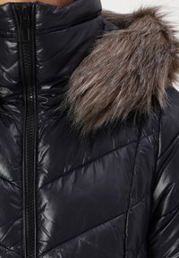 Crna pufasta jakna s visokim ovratnikom, koja ima kapuljaču od umjetnog krzna. Jakna ima sjajnu teksturu i kvadratni quilted uzorak s prednjim patentnim zatvaračem.