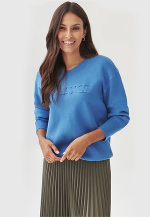 Blaues Sweatshirt mit geprägtem "BALANCE"-Text, rundem Ausschnitt und gerippten Bündchen, kombiniert mit einem gefalteten olivgrünen Rock.