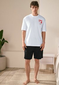 White short-sleeve T-shirt with a red graphic design on the chest, paired with black shorts. Material appears soft; relaxed fit.