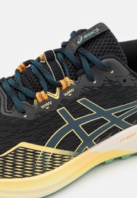 Zapatilla deportiva negra con una parte superior de malla transpirable y detalles en teal y naranja, una suela texturizada y elementos reflectantes.