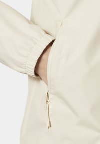 Beige jack met elastische manchetten, een ritszak aan de zijkant met een trekband en een gladde, gestructureerde stof. Focus op functionele ontwerpelementen.