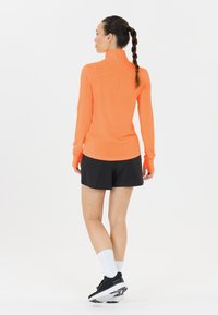 Kvinde med fletninger iført en orange langærmet trøje, sorte shorts, hvide sokker og sorte løbesko, stående imod en hvid baggrund.