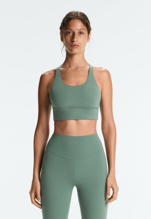 Frau trägt passenden grünen Sport-BH und hoch taillierte Leggings, steht vor einem schlichten weißen Hintergrund mit neutralem Gesichtsausdruck.
