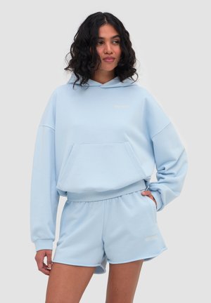 Frau mit lockigem dunklem Haar, die einen hellblauen oversized Hoodie und passende Shorts trägt und mit einer Hand in der Tasche vor einem einfarbigen Hintergrund steht.