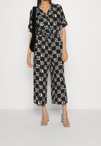 Combinaison à motifs noir et blanc avec manches courtes, taille nouée et coupe jambes larges. Associée à des sandales à talons hauts à carreaux.