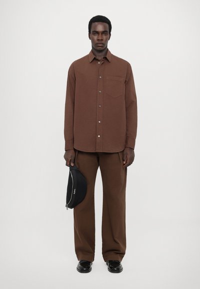 Chemise brune à boutons avec une poche, assortie à un pantalon brun. Tient un sac accessoire noir. Design simple et épuré.