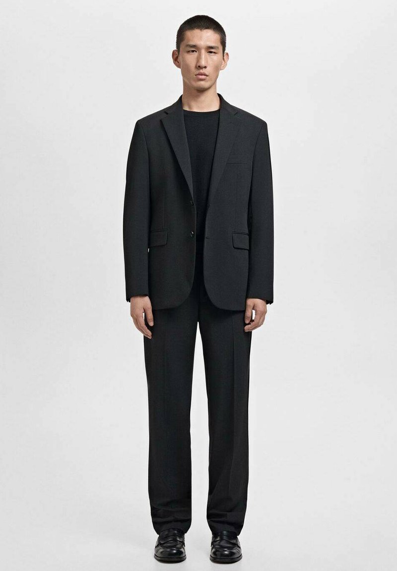 Traje negro con un blazer ajustado y solapas con muesca, combinado con pantalones rectos, llevado sobre una camisa negra y zapatos.