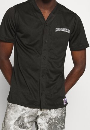 Mann trägt ein kurzärmeliges schwarzes Sporthemd mit Knöpfen und dem Schriftzug "LOS ANGELES" sowie grau gemusterte Shorts, die Arme entspannt hinter dem Rücken.