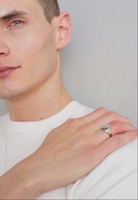Anello d'argento con una pietra nera, indossato su una mano appoggiata su una spalla. Lo sfondo è grigio chiaro; la persona indossa una maglietta bianca a coste.
