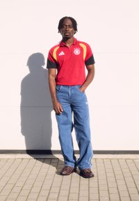 Κόκκινο και μαύρο polo shirt Adidas με λογότυπο Γερμανίας, μπλε τζιν παντελόνι και σκούρες καφέ αθλητικές παπούτσια, στέκεται μπροστά σε ανοιχτόχρωμο τοίχο.