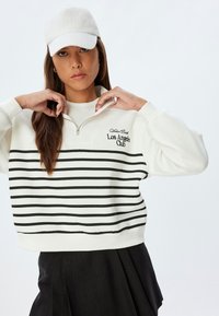 Sweater putih yang dipotong dengan garis horizontal hitam, dilengkapi dengan kerah setengah resleting dan teks "Los Angeles Club." Ditemani dengan topi yang serasi.