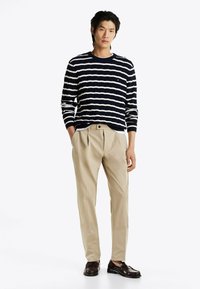 Navy- und weiß gestreifter Strickpullover, beigem Plissee-Hosen, dunkle Mokassins. Der Pullover hat einen gerippten Halsausschnitt; die Hosen besitzen seitliche Taschen.