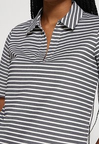 Femme portant un polo rayé horizontal noir et blanc avec col et manches courtes, cheveux coiffés en longues tresses.