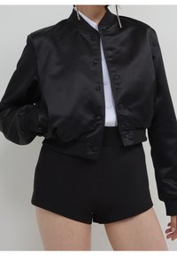 Veste bomber courte en satin noir avec un col montant et une fermeture à boutons, associée à un short noir taille haute. Texture lisse, design décontracté.