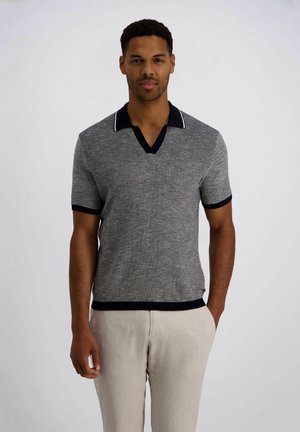 Mann trägt ein graues, kurzärmeliges, gestricktes Poloshirt mit schwarzem Besatz und hellbeige Hosen, steht vor einem einfarbigen Hintergrund.