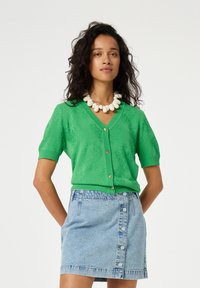 Groene kortebloemige cardigan met gestructureerde fruitpatronen en een V-halslijn, voorzien van lichtgekleurde knopen. Gecombineerd met een spijkerrok.