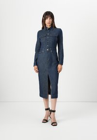 Denimkjole med innsvingt liv, lange ermer, knappestolpe foran, to brystlommer og splitt foran. Mørk blå farge med oransje søm.