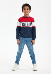 Pull rayé rouge, marine et blanc avec le texte "EASTSIDE", associé à un jean bleu clair et des baskets blanches. Design simple et décontracté.