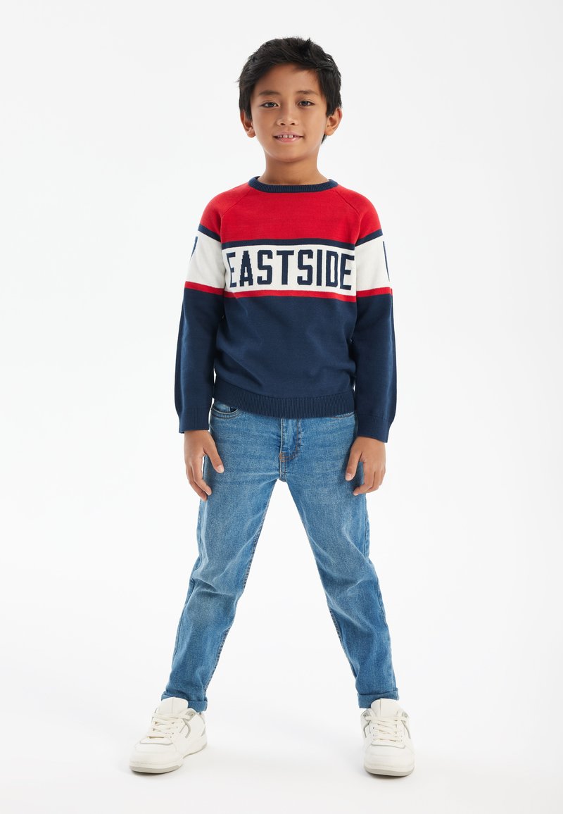 Pull rayé rouge, marine et blanc avec le texte "EASTSIDE", associé à un jean bleu clair et des baskets blanches. Design simple et décontracté.