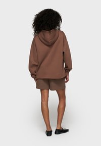 Personne avec des cheveux bouclés portant un sweat à capuche marron et un short assorti, se tenant de dos face à un fond uni.