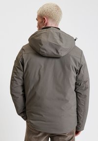 Giacca isolata grigia con cappuccio, caratterizzata da una texture liscia e un design semplice. Dettagli notevoli includono lacci regolabili sul cappuccio.