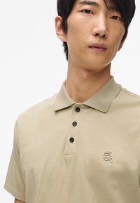 Bežová polo košeľa s golierom, tromi čiernymi gombíkmi a bielym vyšitým logom na hrudi. Mäkká látka a voľný strih.