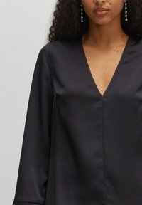 Schwarze Satinbluse mit tiefem V-Ausschnitt, langen Ärmeln und glatter Textur. Subtile Nähte entlang der Vordernaht für zusätzliche Details.