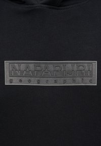 Μαύρο ύφασμα με ορθογώνιο λαστιχένιο έμπλαστρο που φέρει ανάγλυφο κείμενο "NAPAPIJRI geographic" στο κέντρο της επιφάνειας.