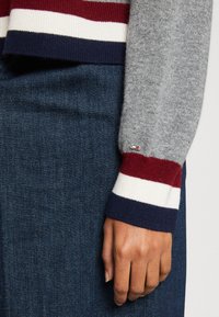 Grauer Pullover mit roten, weißen und marineblauen gestreiften Bündchen; glatte Textur. Dunkelblaue Jeans darunter, auf denen eine Hand am Taillenbund ruht.