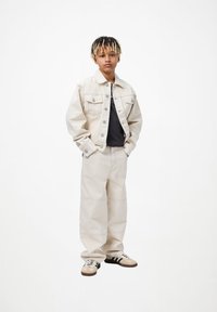 Joven con dreadlocks de puntas rubias que lleva una chaqueta de mezclilla beige y pantalones beige, camiseta negra y zapatillas beige con rayas negras.