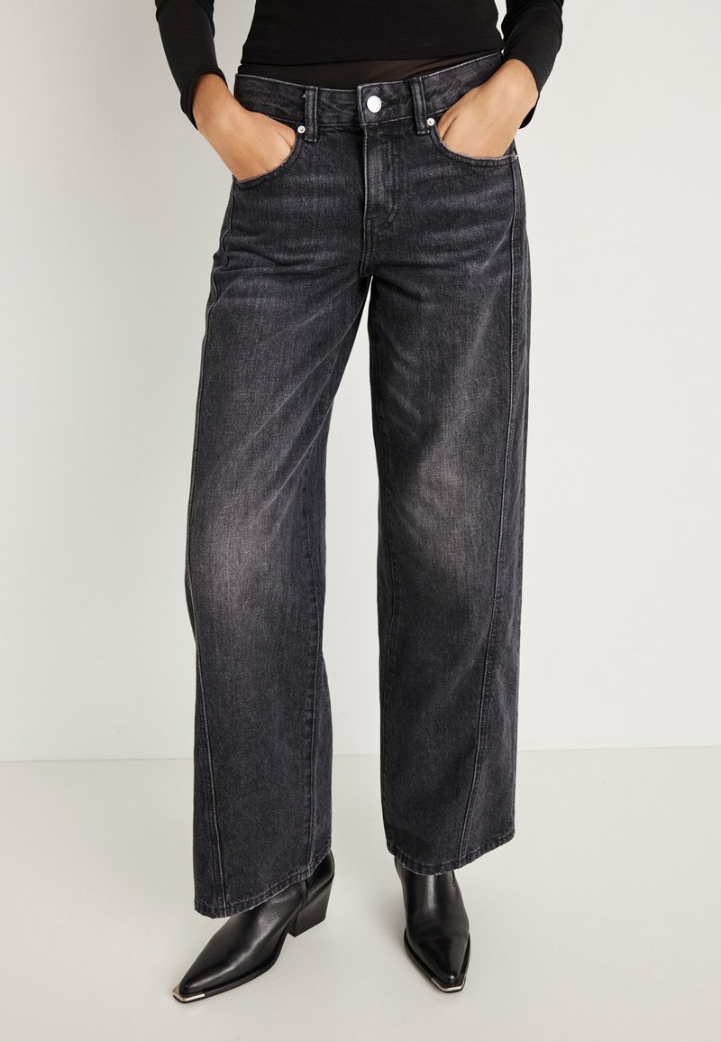 Žena v čiernych vyblednutých širokých džínsoch, čiernych kožených členkových čižmách s ostrou špičkou a čiernom dlhom rukáve s rukami v kapse.