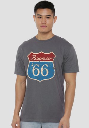 Young man wearing a gray t-shirt with a vintage red and blue shield emblem reading "Bronco '66" on the front.