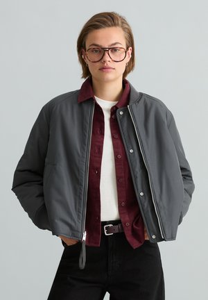 Junge Person trägt übergroße graue Bomberjacke über burgunderfarbenem Hemd und weißem Top, mit kurzem Haar und großen braunen Brillen, Hände in den Taschen.