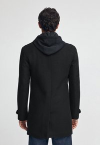 Svart kappa med texturerad yta, med huva och slim fit. Synliga manschetter, slits bak och en mjuk innervadding.