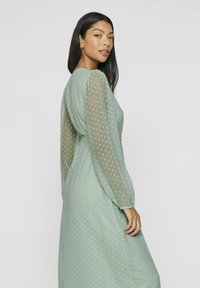 Grønn prikkete kjole med gjennomsiktige lange ermer og belted midje. Stoffet har en myk, jevn tekstur som gir en flytende silhuett.