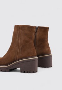 Botas de tobillo de ante marrón con una plataforma gruesa y una suela texturizada, que cuentan con un cierre lateral y detalles de costura a lo largo de las costuras.