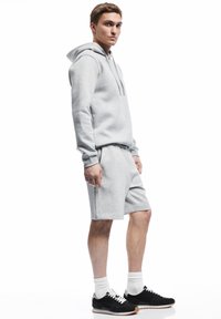 Sudadera gris con capucha y pantalones cortos a juego, ambos en una suave mezcla de algodón. Zapatillas negras, calcetines blancos y un diseño casual típico del athleisure.