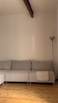 Ανοιχτό γκρι γωνιακός καναπές με διπλωμένη μπεζ κουβέρτα στη δεξιά πλευρά, ξύλινο πάτωμα, ψηλό ασημένιο επιδαπέδιο φωτιστικό και μονόχρωμος μπεζ τοίχος με σκούρα δοκό από πάνω.