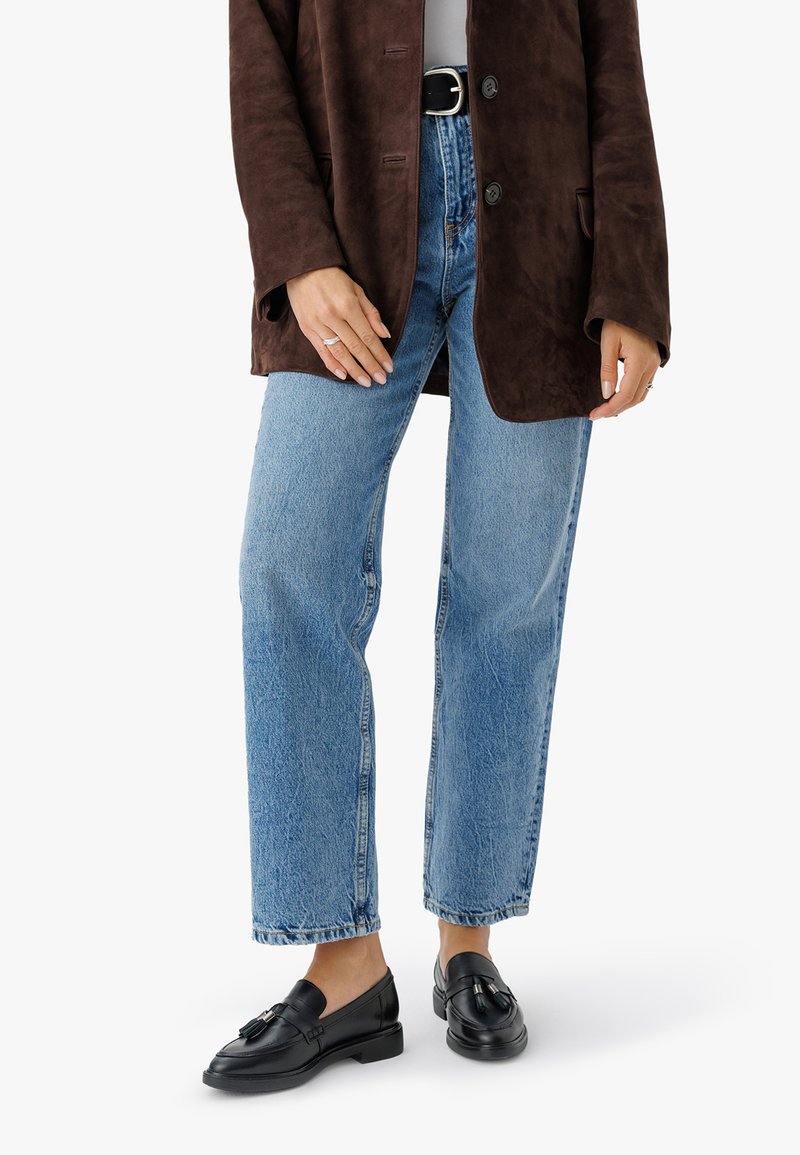 Kvinna i ljusblå jeans, svarta läderloafers med tofsar, en brun mockajacka och ett svart bälte med en silverspänne.