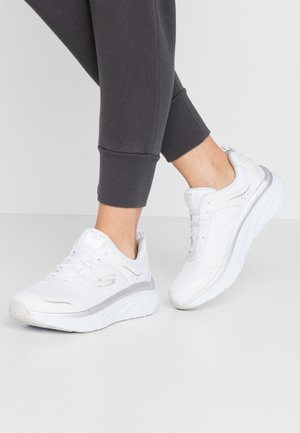 Person iført hvide Skechers sportssko og mørkegrå joggebukser står på en lys gulv mod en ensfarvet baggrund.