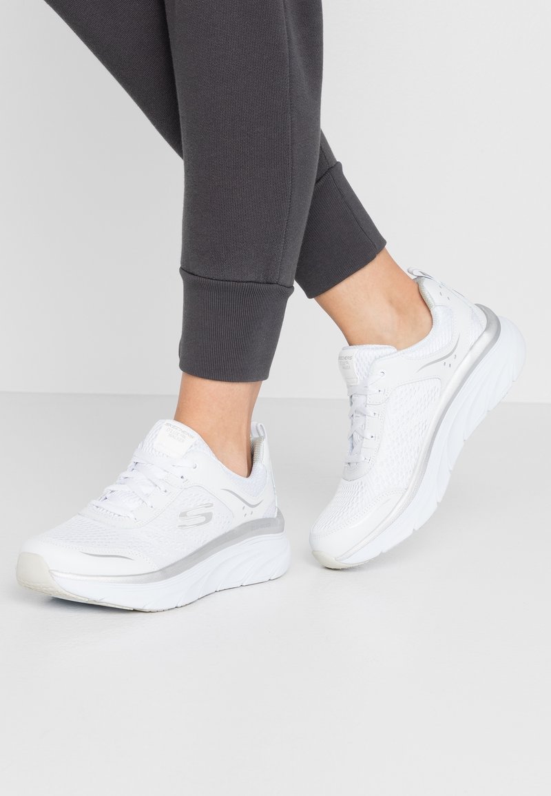 Personne portant des chaussures de sport blanches Skechers et un pantalon de jogging gris foncé, debout sur un sol clair devant un fond uni.