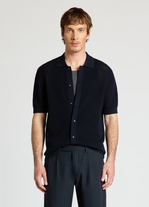 Homme aux cheveux courts et bruns portant un cardigan noir à manches courtes boutonné et un pantalon foncé, debout devant un fond clair uni.