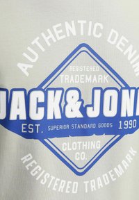 Graues T-Shirt mit blauer und weißer Grafikschrift, die "JACK & JONES" und "AUTHENTIC DENIM" zeigt. Rundes Design, glatte Textur, lässiger Stil.
