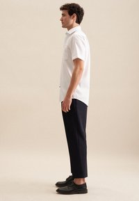 Witte T-shirt met korte mouwen en een borstzak, gecombineerd met marineblauwe tapered broek en zwarte veterschoenen, zijaanzicht.