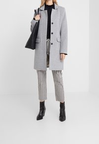 Grahamfarbener Wollmischungsmantel mit Reverskragen und vorderem Knopfverschluss, kombiniert mit schwarz-weiß gestreiften Hosen und schwarzen Ankle Boots.