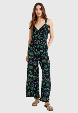 Kobieta stoi w czarnym bezrękawnikowym kombinezonie w zielono-niebieski kwiatowy wzór i złotych sandałach na jednolitym jasnym tle.